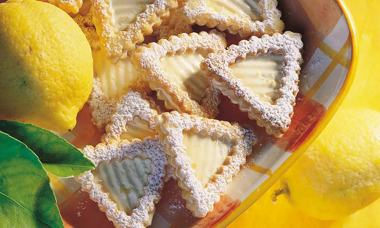 Dreieckige Kekse mit gezacktem Rand, gefüllt mit gelber Zitronencreme und bestäubt mit Puderzucker, angerichtet auf bunter Schale