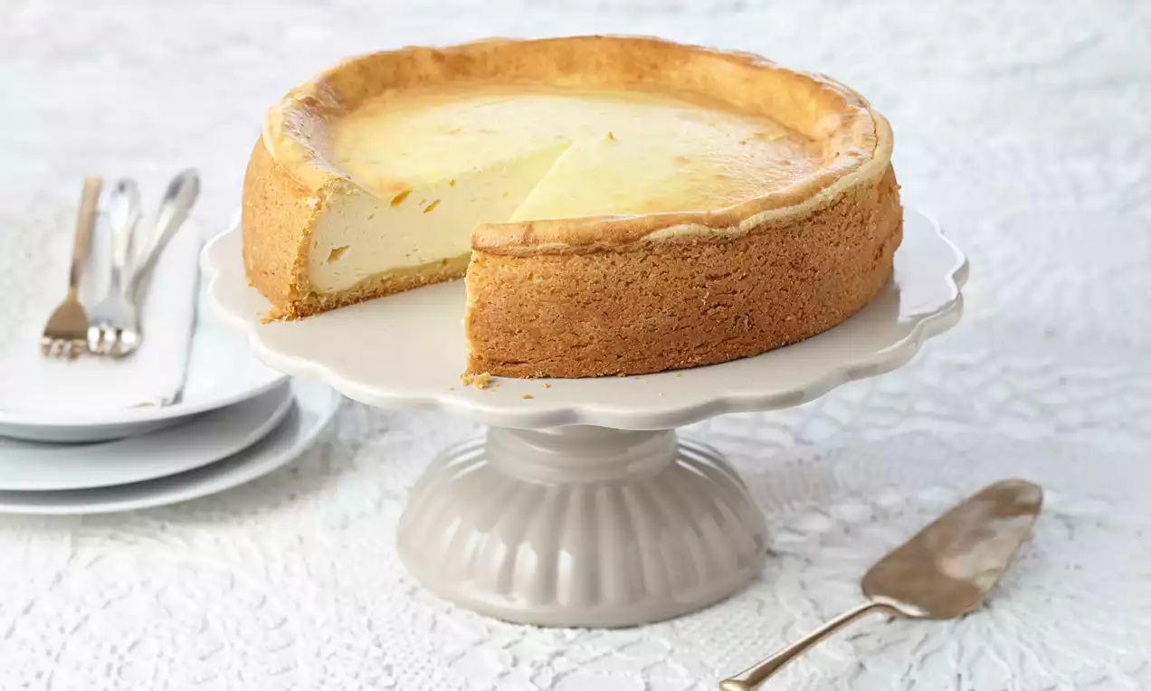 Goldbrauner Käsekuchen mit glatter, hellgelber Füllung auf weißer Tortenplatte, angerichtet neben Besteck und Teller auf Spitzenunterlage