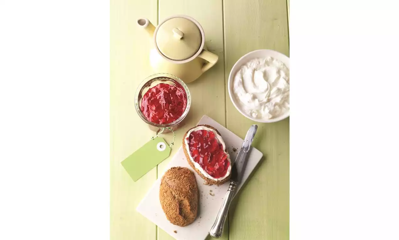Červený ríbezľový džem v skle vedľa hnedého pečiva s džemom a šľahačkou na bielom tanieri, doplnené žltou kanvicou na zelenom stole