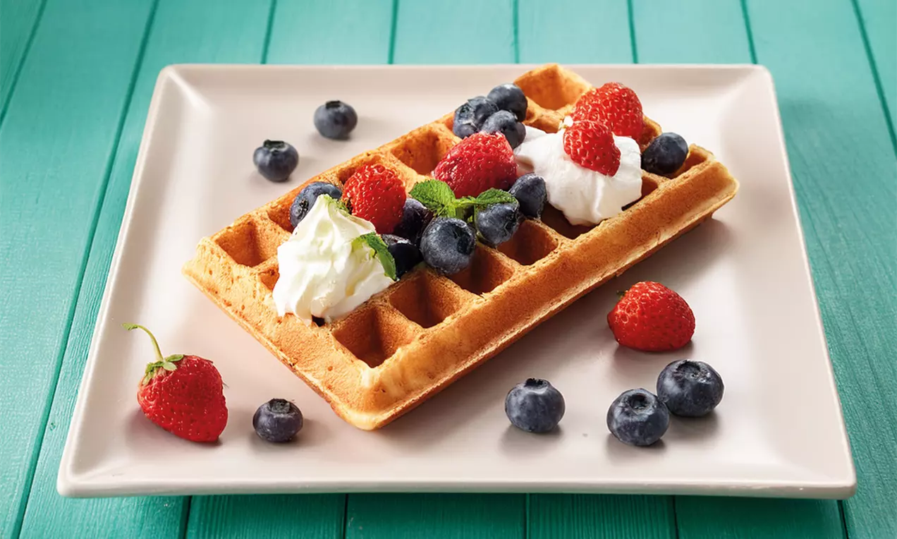 Goudbruine rechthoekige wafel met luchtige slagroom, rode aardbeien, blauwe bessen en een groen blaadje op een wit vierkant bord