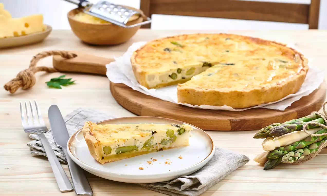 Goldbraune Spargelquiche mit cremiger Füllung und grünen Spargelstücken, serviert auf einem Holzbrett neben einer Portion auf Teller