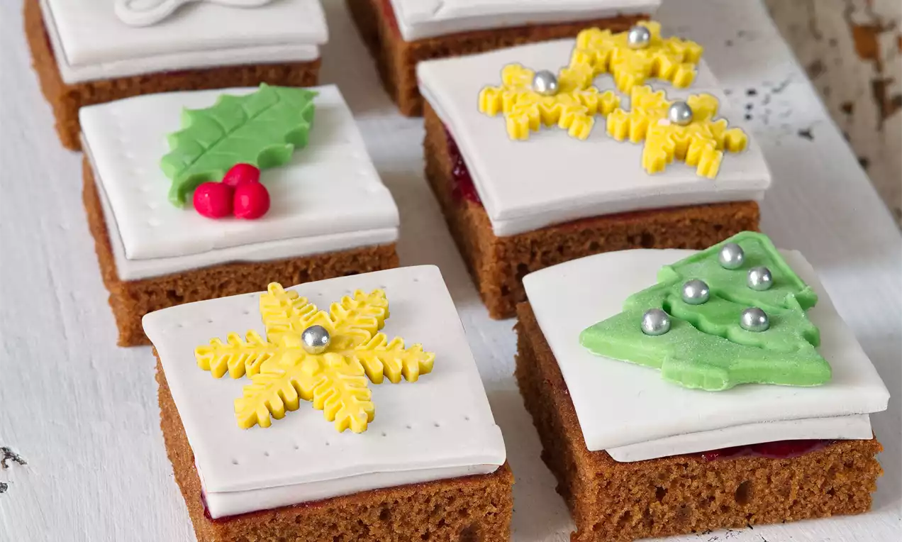 Čtvercové perníčky s bílou cukrovou polevou zdobené barevnými motivy listů, hvězd a stromečků, uspořádané na světlém podkladu