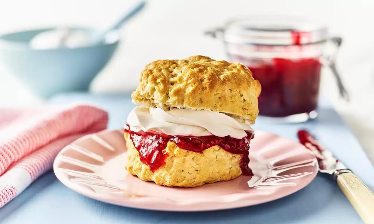 Golden vegan scone with a craggy top, filled with glossy red jam and fluffy cream, on a pastel pink plate with a jam jar nearby
