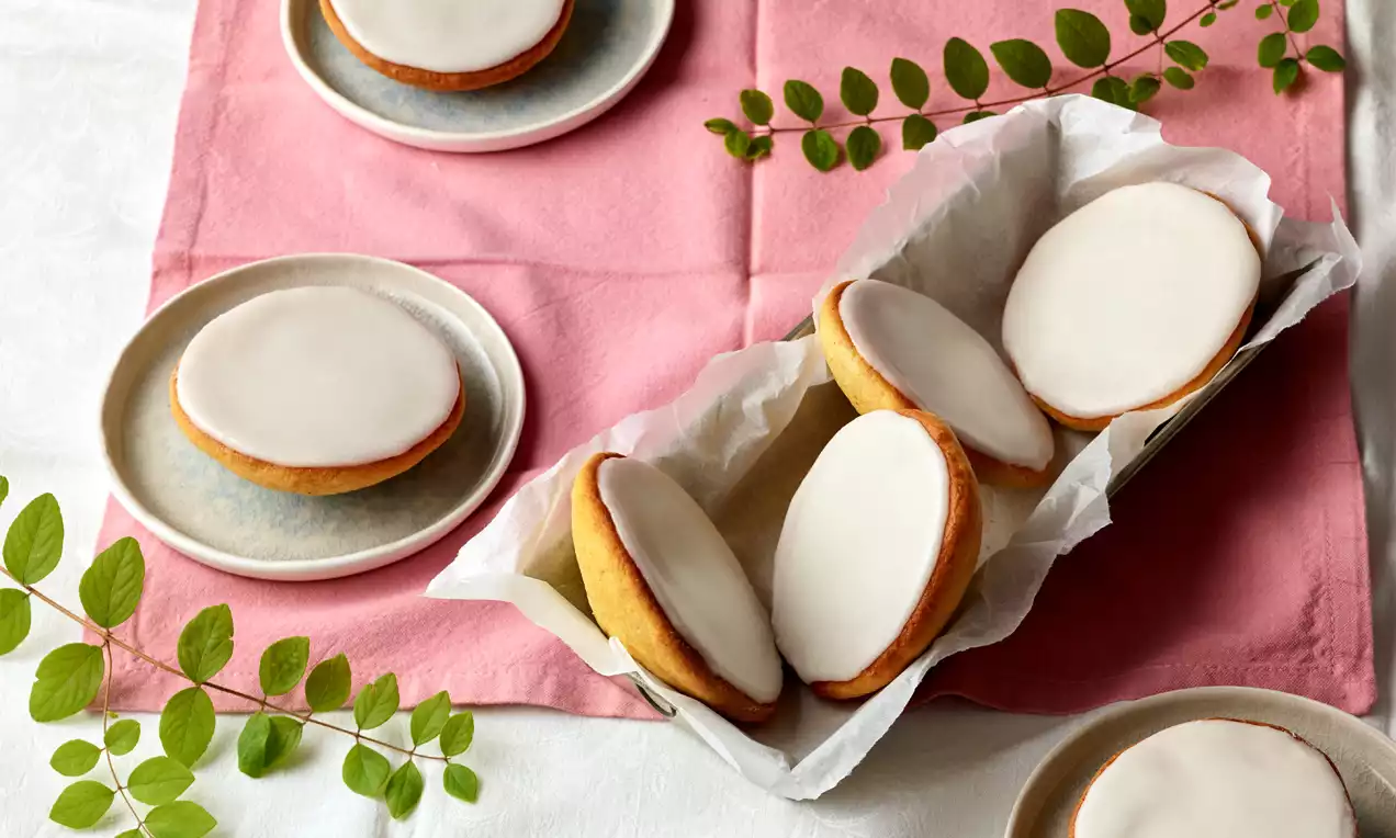 Kleine goldbraune Amerikaner mit glatter weißer Zuckerglasur, teilweise auf Tellern und in einer Schale mit Backpapier arrangiert