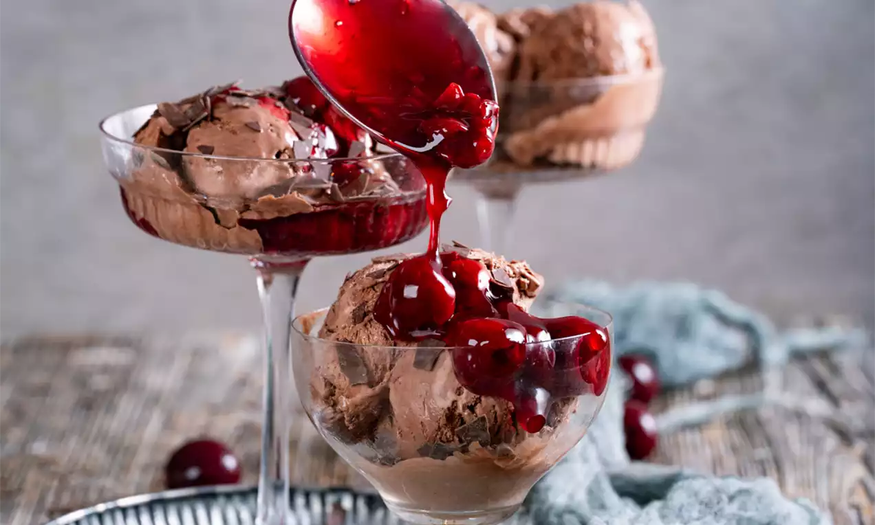 Temno rjav čokoladni sladoled v steklenih čašah, prelit z gostim rdečim višnjevim prelivom in okrašen s sočnimi celimi višnjami