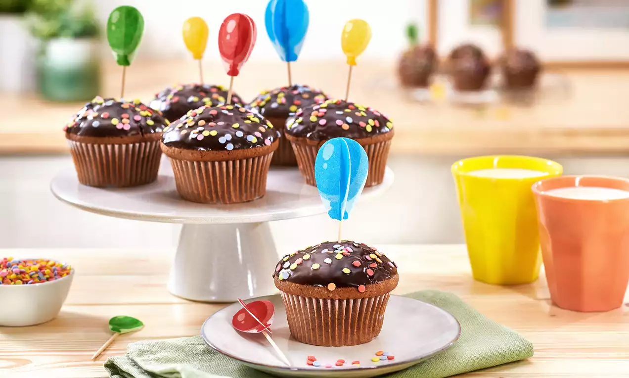 Schokoladenmuffins mit bunten Zuckerstreuseln und Luftballonförmigen Picks, arrangiert auf Teller und weißer Etagere
