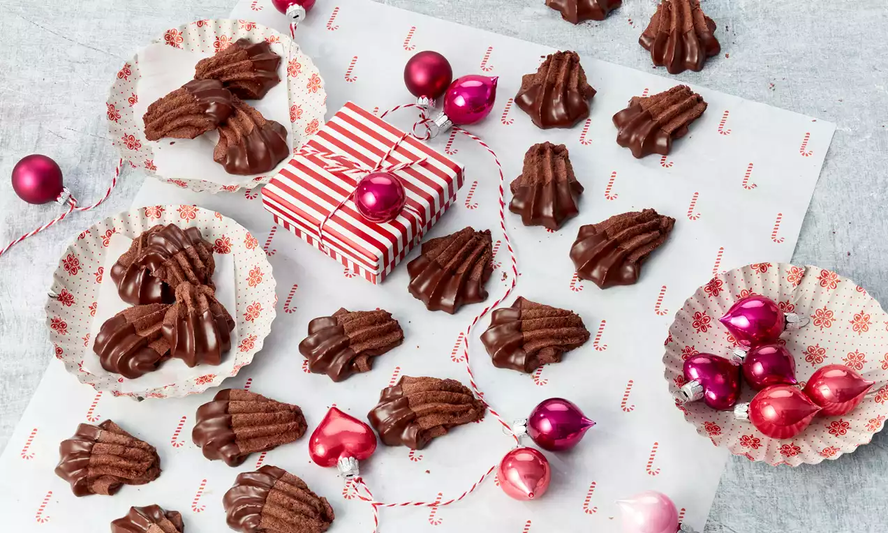 Tamnosmeđi keksi rebraste površine u obliku otisaka šapa, složeni na papir s blagdanskim uzorkom, uz ukrasne kuglice i poklon kutiju