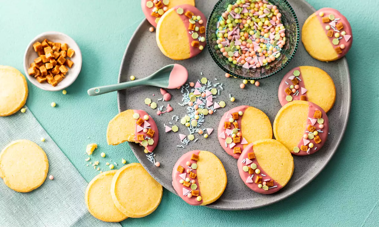 Okrugli keksi zlatnožute boje prekriveni glazurom roze nijanse s raznobojnim šarenim posipom na sivom tanjuru i plavkastoj podlozi