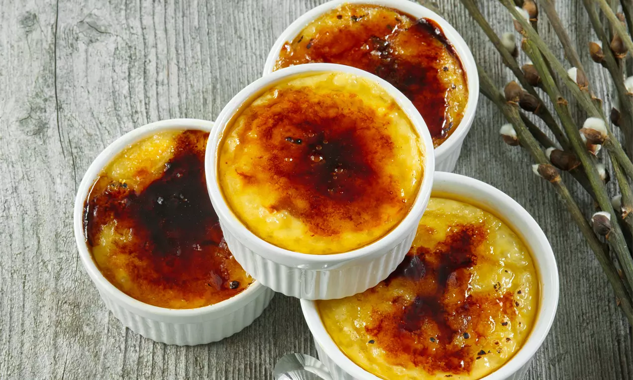 Zlatisté tapiokové dezerty s karamelovou krustou v bielych keramických mištičkách, uložené na drevenom povrchu vedľa vŕbových konárikov