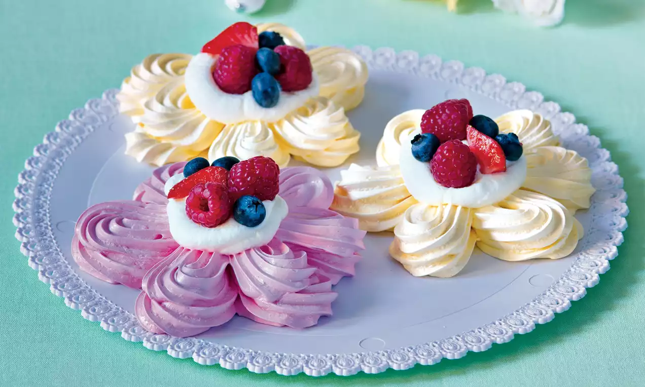 Barevné květinové pavlovy s jemnými meringue lístky, bílým krémem uprostřed a čerstvými malinami, borůvkami na dekorativním talíři