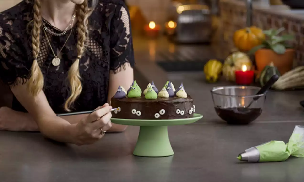 A round chocolate cake on a pale green stand, topped with colorful buttercream kisses and candy eyes, set on a counter with Halloween decor