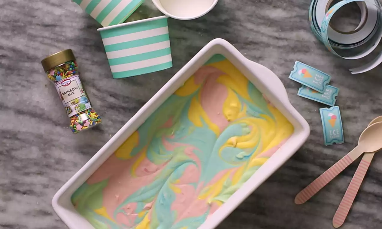 Regnbuefarvet is med bløde pastelfarver i gul, lyserød og blå, hvirvlet sammen i et hvidt rektangulært fad på en grå marmorbordplade