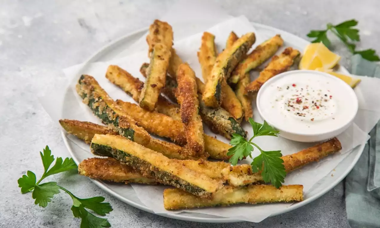 Golden fried zucchini straws with a crispy breadcrumb coating arranged on a plate with creamy dip, parsley garnish, and lemon wedges