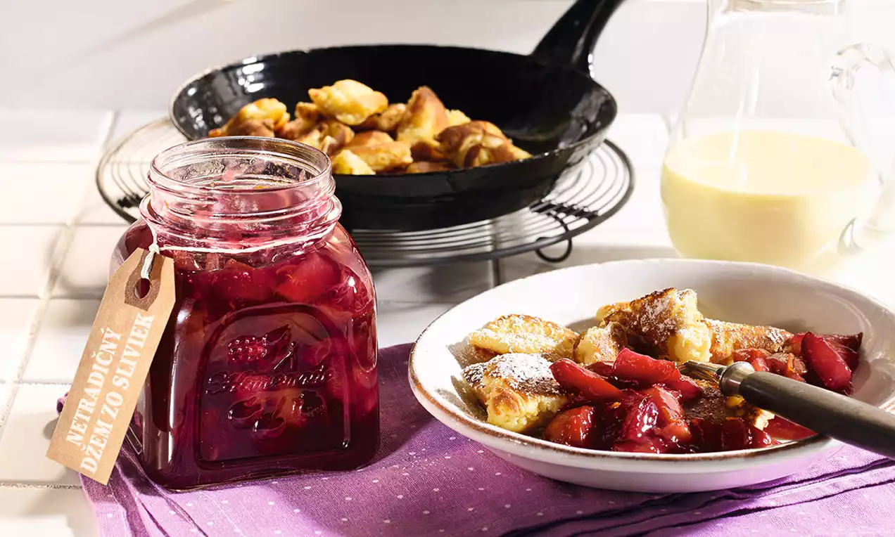 Sklenený pohár s tmavočerveným slivkovým džemom vedľa taniera s opečenými kúsokmi cesta a kúskami ovocia, na fialovej utierke