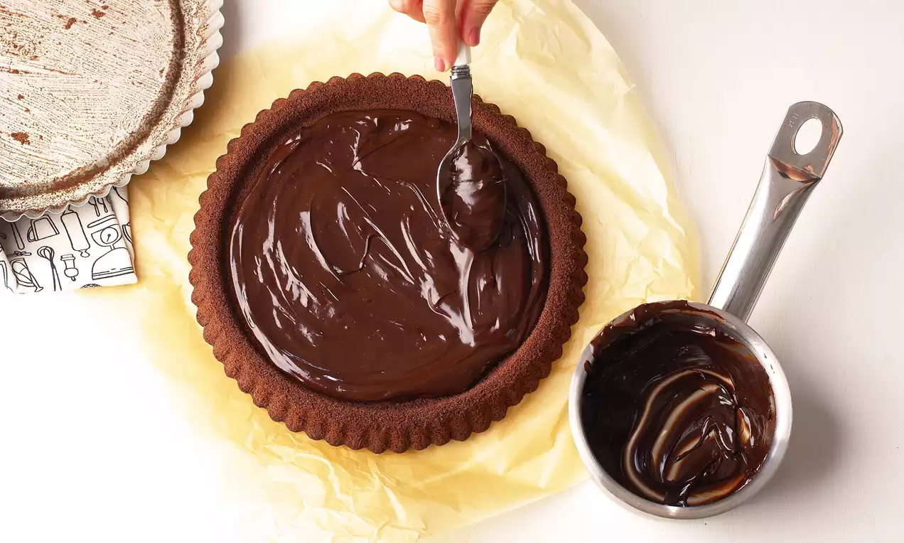 Kakao rengi tart kalıbı içinde yoğun ve pürüzsüz çikolata kreması, yanında metal kapta kalan krema ve şeffaf kağıt üzerinde sunulmuş