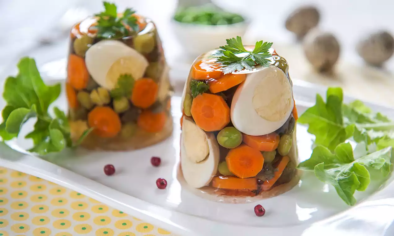 Mini galaretki w formie półprzezroczystych stożków z jajami przepiórczymi, marchewką, groszkiem i natką, na białym talerzu z sałatą i różowym pieprzem