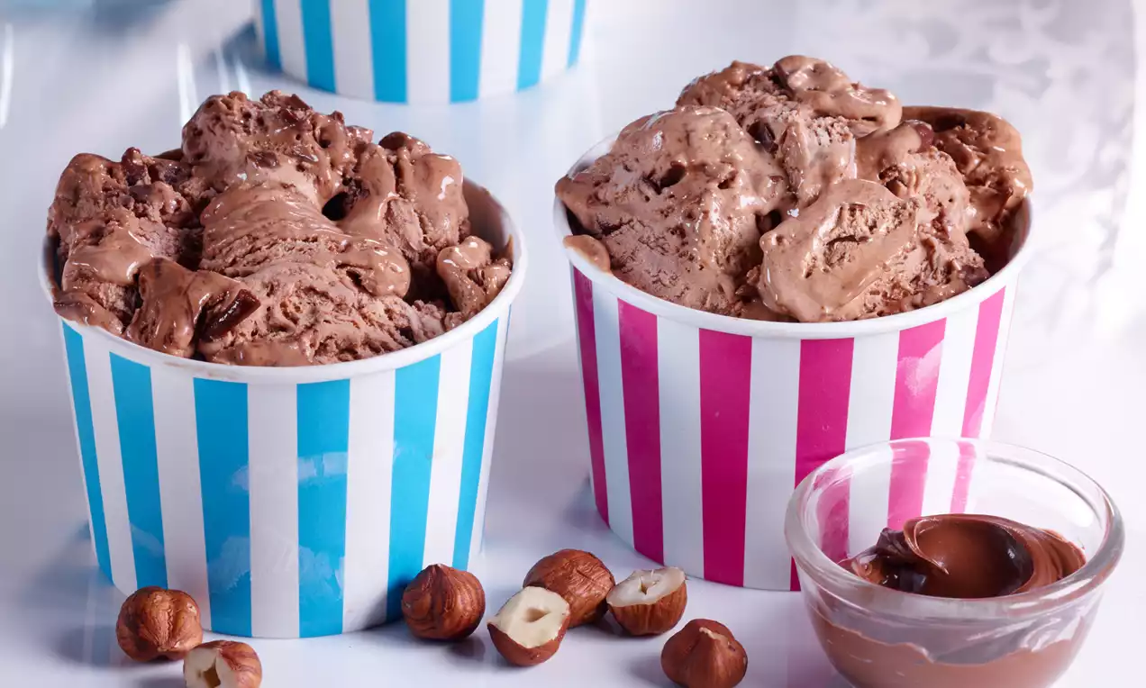 Cremiges schokoladenbraunes Nutella-Eis mit leicht glatter Textur in gestreiften Bechern, garniert mit Haselnüssen und einer kleinen Schale Nutella