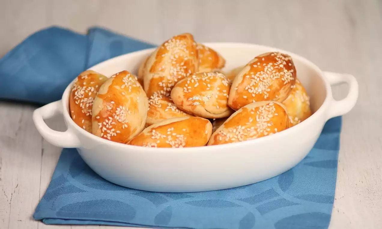 Altın rengi, susam serpilmiş, hafif kabarmış tuzlu poğaçalar beyaz kulplu bir kapta, mavi bir örtü üzerinde düzenlenmiş