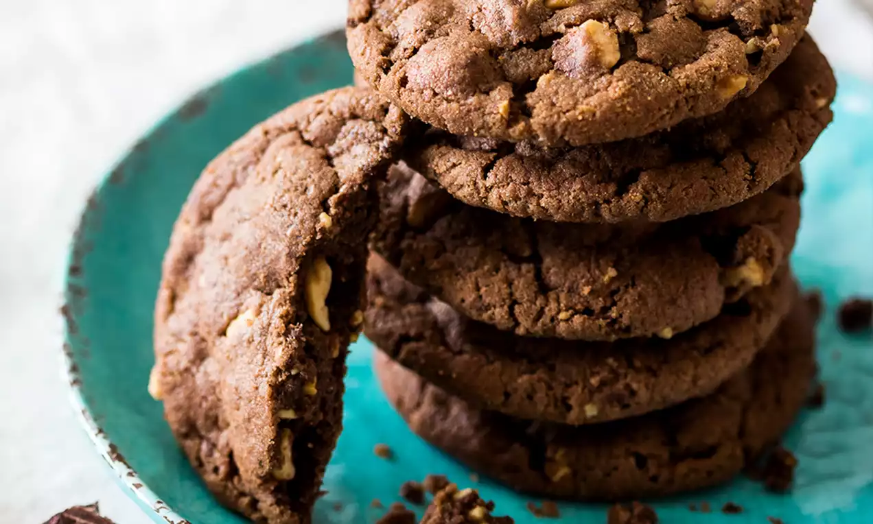 Cookies tondi color cacao con punte di frutta e pezzi di frutta secca dorati, disposti a pila su un piatto azzurro con dettagli vintage