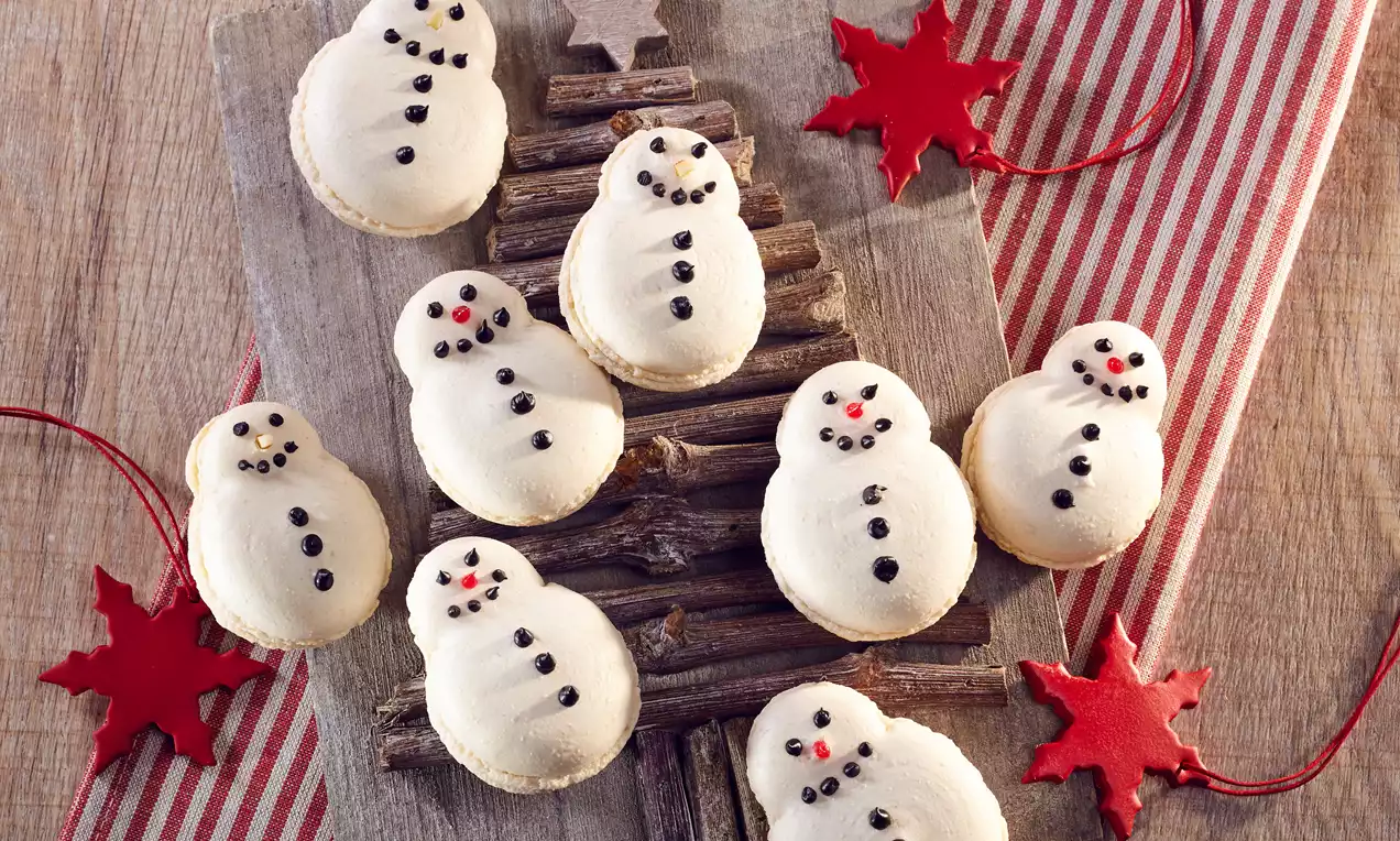 Kardan adam şeklinde beyaz makaronlar, çikolata ve kırmızı detaylarla süslenmiş, ahşap bir zemin ve kırmızı yıldızlarla çevrili