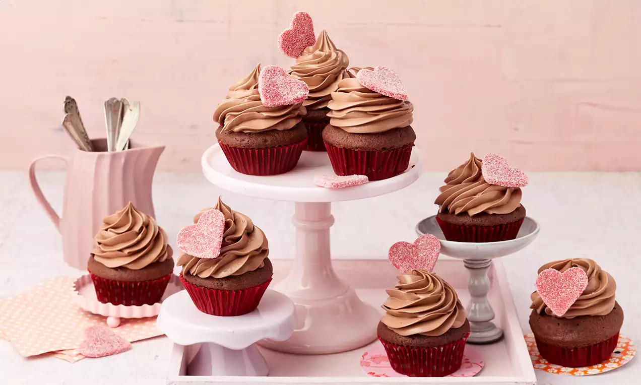 Schokoladen-Cupcakes mit cremig brauner Sahnehaube und rosa Zuckerdekor-Herzen auf roten Förmchen, arrangiert auf rosa Etageren