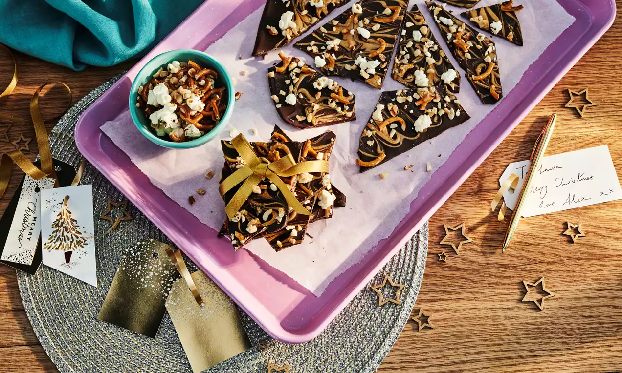 Dark shards of glossy chocolate topped with tangled pretzels, nuts, and popcorn, arranged on pink parchment in a lilac tray