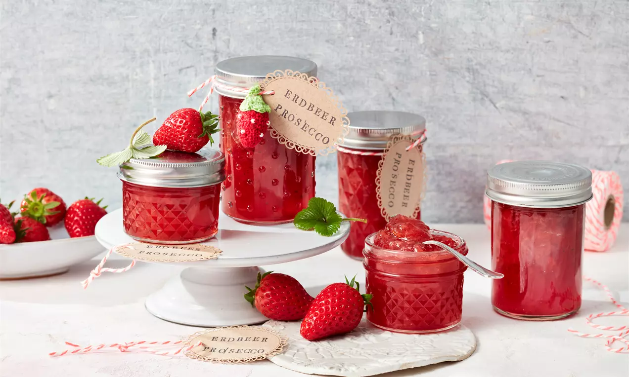 Erdbeer-Prosecco-Marmelade im Glas mit leuchtend roter Fruchtmasse, halbierten Erdbeeren, Minzblatt und Holzlöffel auf einem Brett