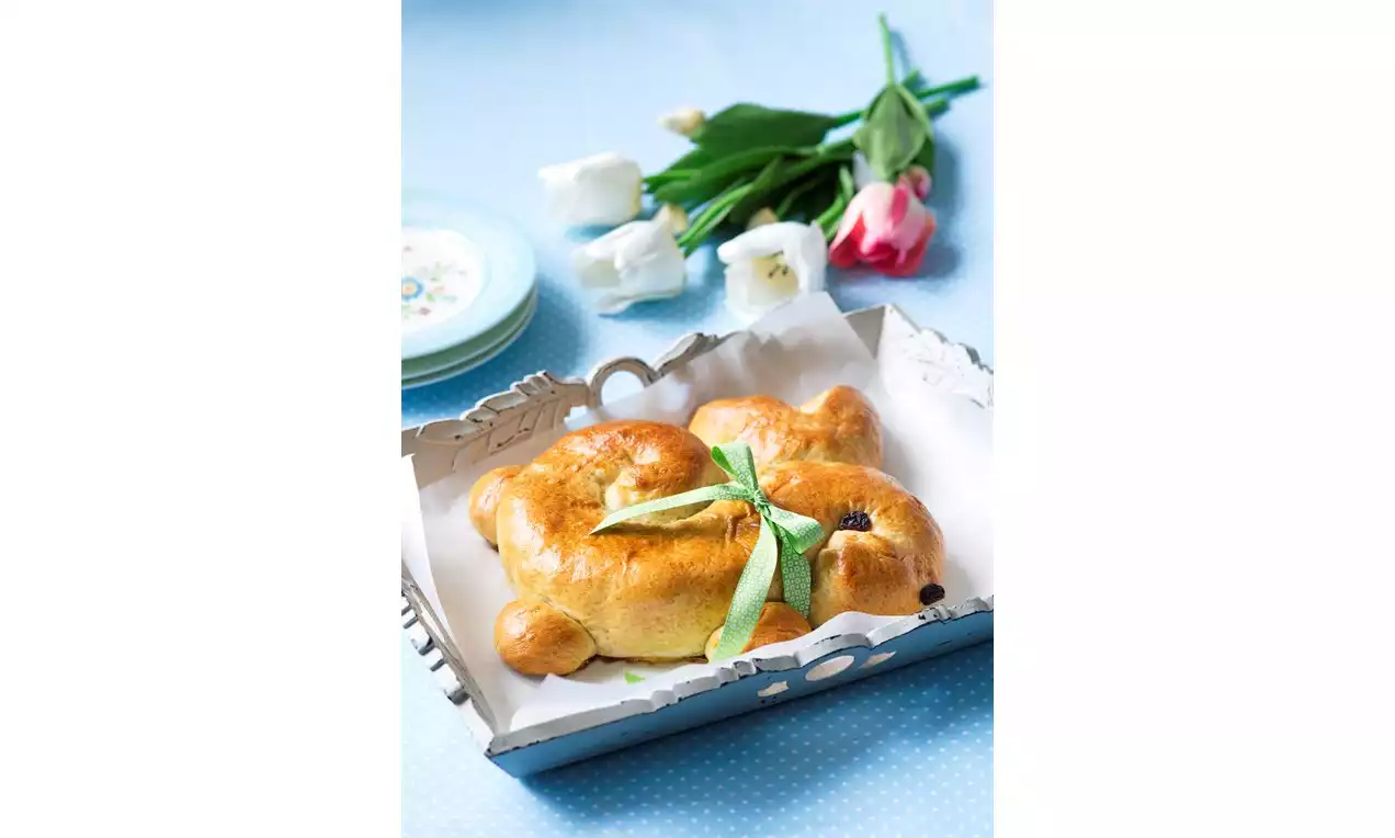 Velikonoční zajíček zlatavé barvy, lesklý povrch, s rozinkovými očky, ozdobený zelenou stuhou, na papíru v kovovém podnosu na světle modrém ubrusu