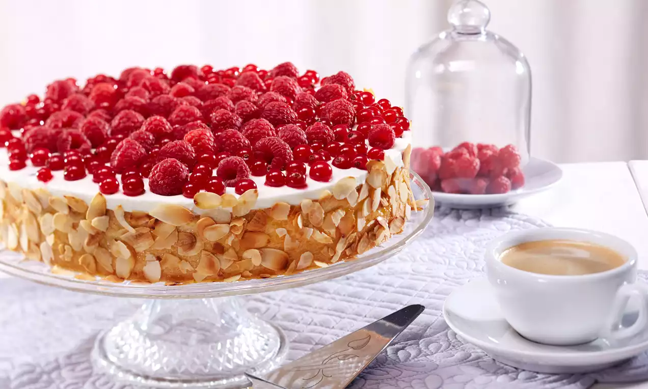 Beeren-Joghurt-Quark-Torte mit glatter weißer Creme, goldbraunen Mandelblättchen und einer leuchtend roten Beeren-Schicht auf Glasplatte