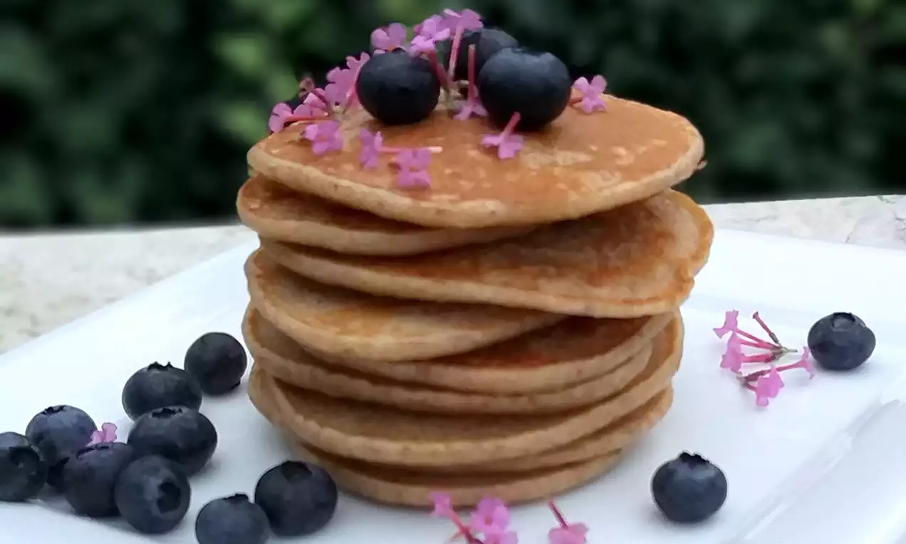 Špaldové lívance vo svetlohnedých odtieňoch na sebe naukladané do veže, ozdobené čučoriedkami a ružovými kvietkami na bielom tanieri