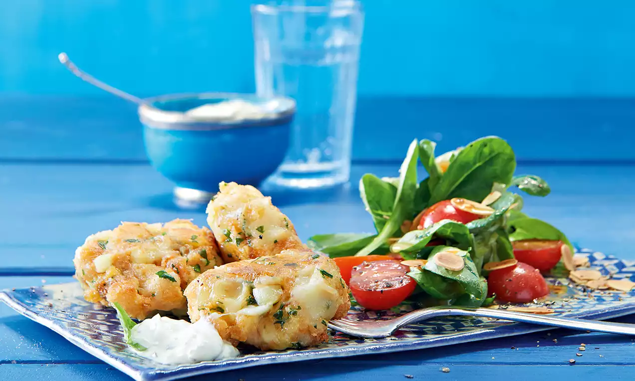 Goldbraune Kaspressknödel mit knuspriger Struktur neben Feldsalat, roten Cherrytomaten und gerösteten Mandelblättern auf einem blauen Teller