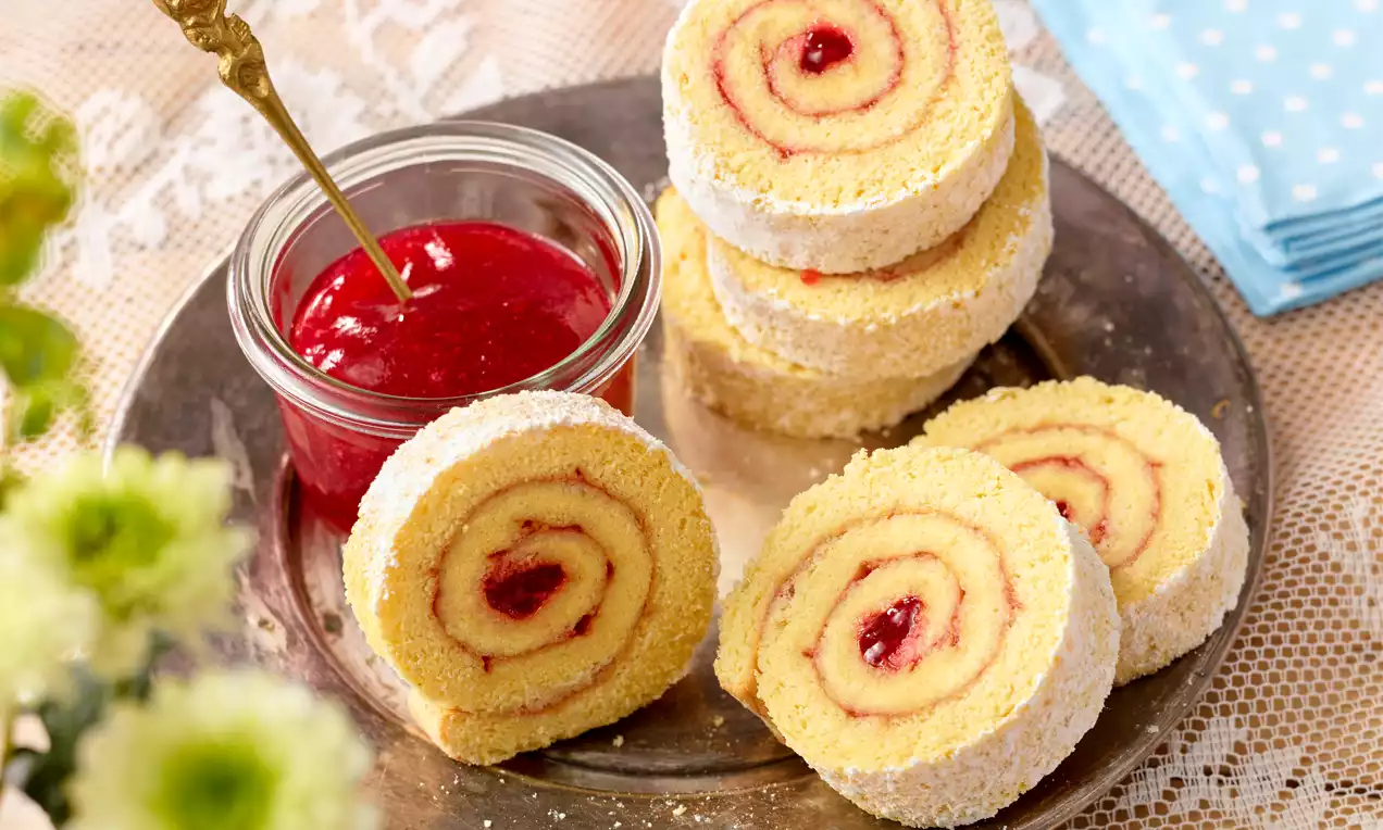 Helle Biskuitroulade in Scheiben geschnitten mit roter Marmeladenfüllung, bestäubt mit Puderzucker, auf einer Platte neben Glas mit Marmelade