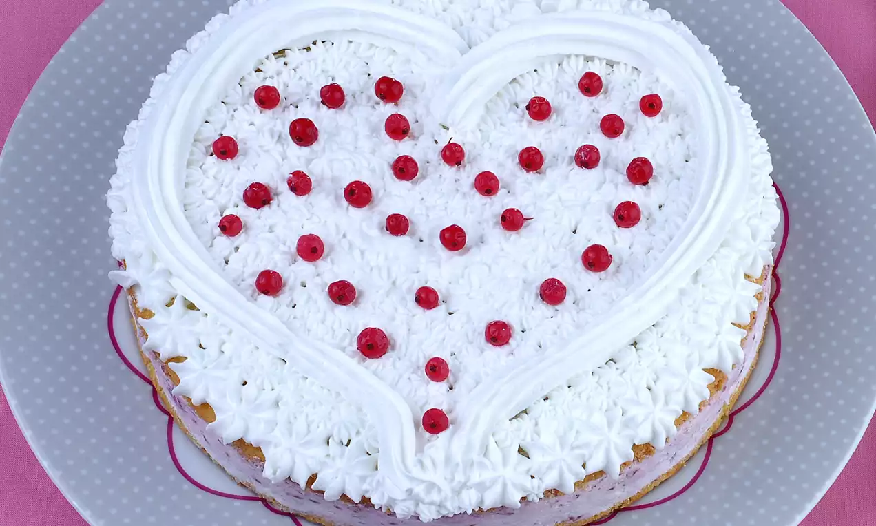 Kalp şeklinde beyaz kremalı pasta, üzerine ahududular serpilmiş ve pembe zemin üzerinde gri puantiyeli tabakta sunulmuş