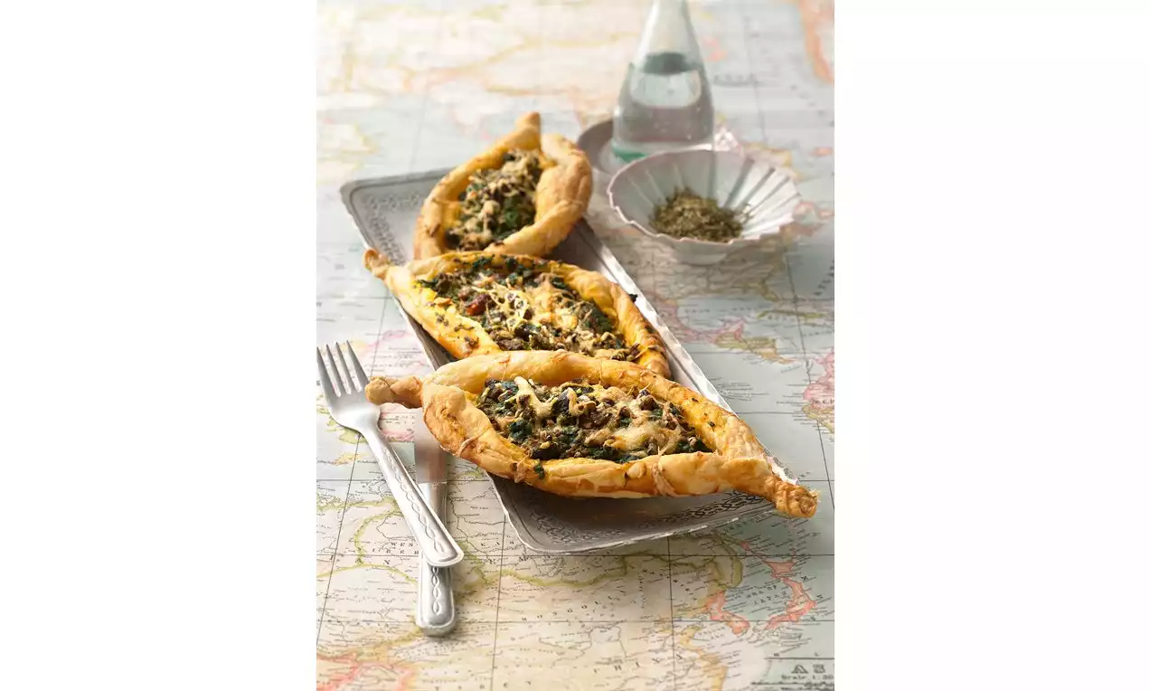 Pečené lodě zlatavého těsta plněné masovo-špenátovou náplní, posypané sýrem, servírované na plechovém tácku na podkladu s mapou