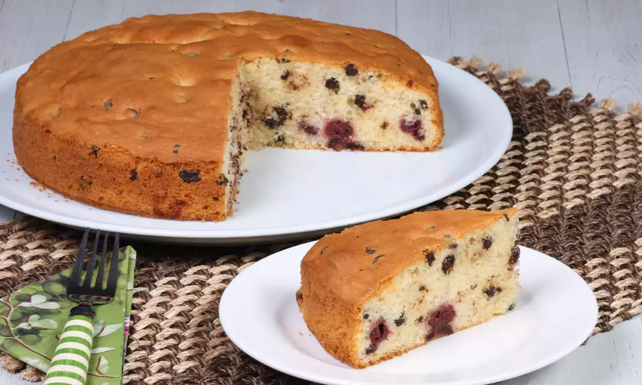 Yuvarlak beyaz tabakta servis edilen, içi vişne ve çikolata parçalı dilimi kesilmiş açık kahverengi kek, dokulu hasır örtü üzerinde