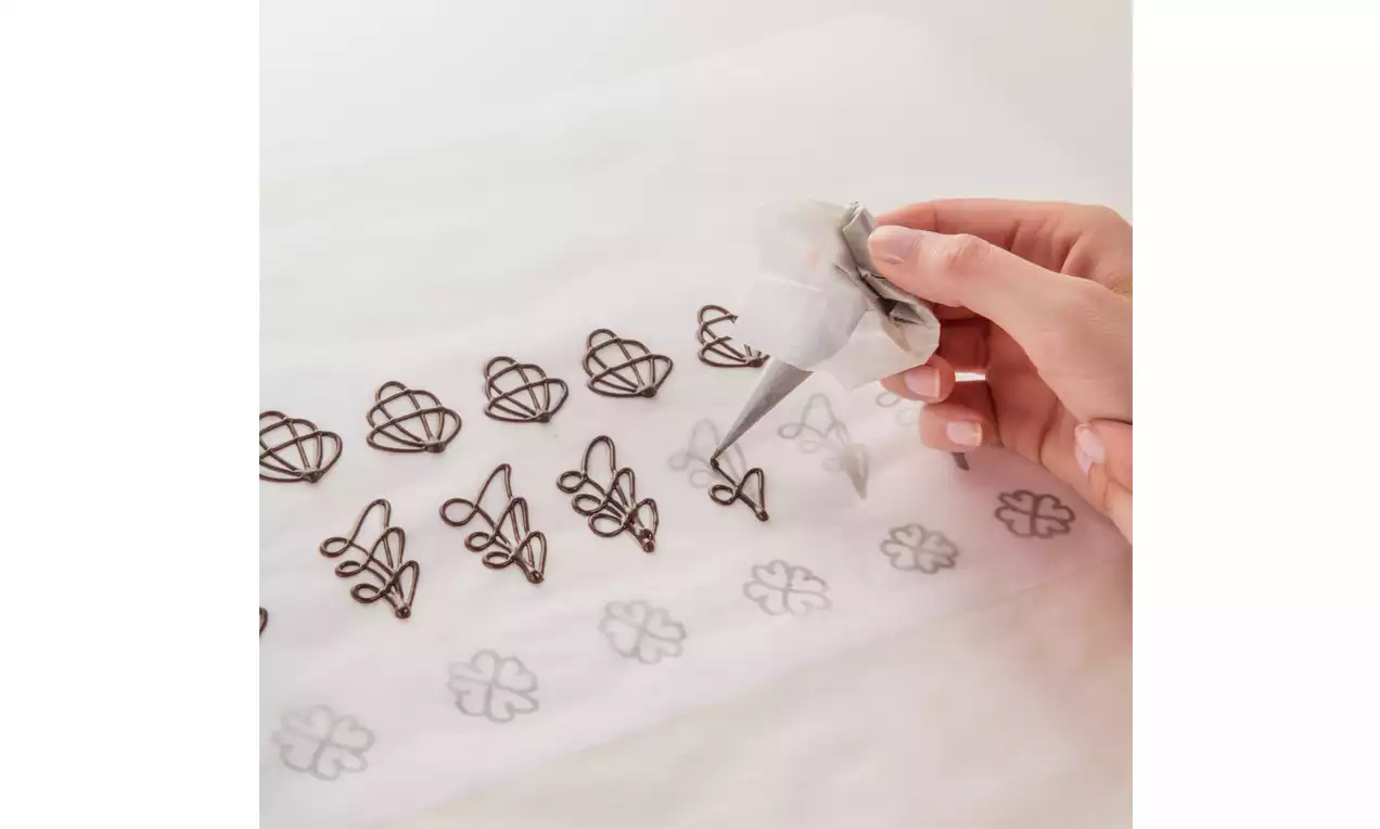Mehrere filigrane Schokoladenornamente in dunkler Farbe auf weißem Backpapier, eine Hand formt Symbole mit einer feinen Spritztülle