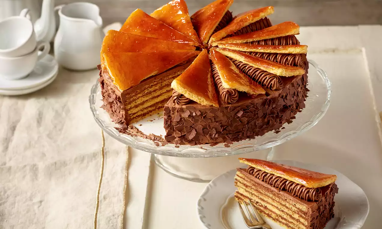 Dobostorte mit glänzenden Karamellspitzen und Schokoladencremeschichten auf Glasplatte, neben Stück auf weißem Teller mit Gabel drapiert