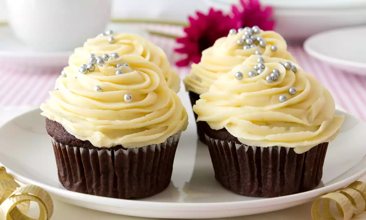 Chocolate cupcakes topped with smooth pale yellow buttercream swirls and silver sprinkles on a white plate with pink flowers in the background