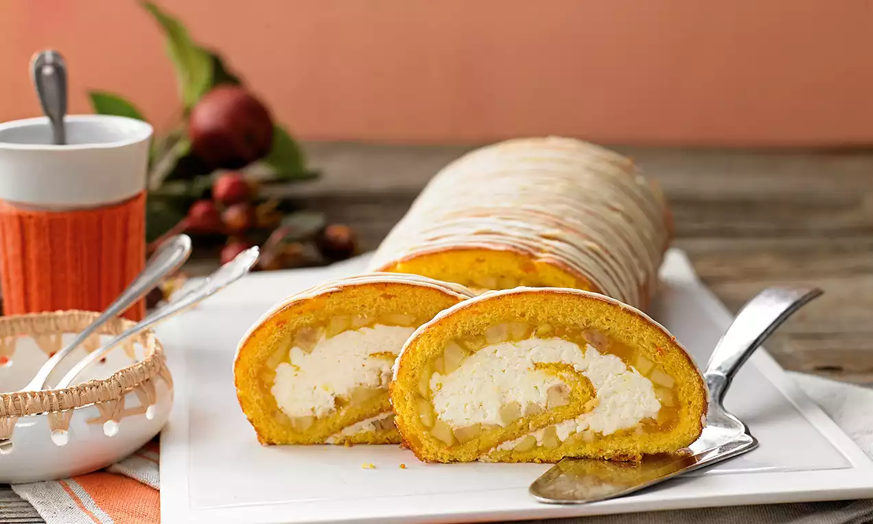 Rolada intenzivno narančaste boje s mekanim nadjevom od bijele kreme, servirana na bijelom pladnju uz zlatnu žlicu i jesensku dekoraciju