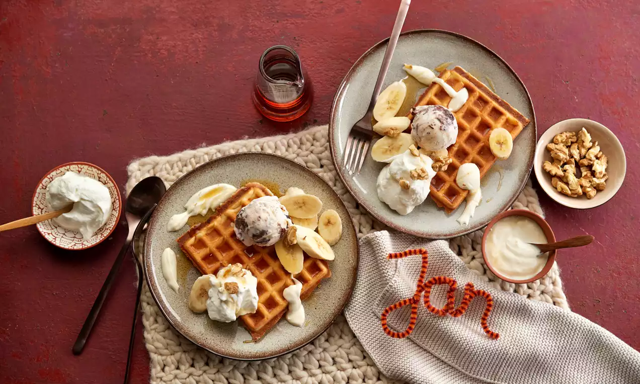 Goldbraune, rechteckige Waffeln mit fluffiger Textur, garniert mit Bananenscheiben, Eis und Sahne auf einem grauen Teller mit Walnüssen
