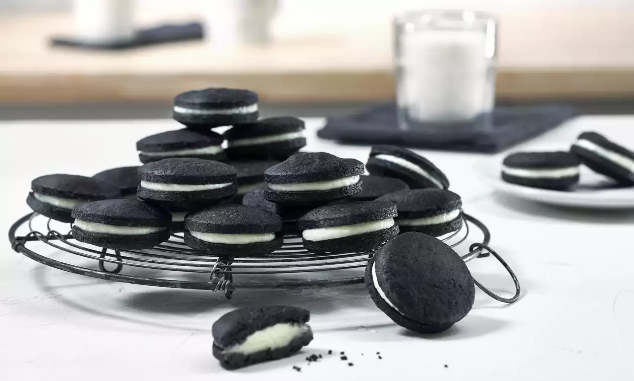 Schwarze Doppelkekse mit weißer Füllung auf einem runden Metallgitter, daneben ein angebissenes Exemplar und ein Glas Milch im Hintergrund