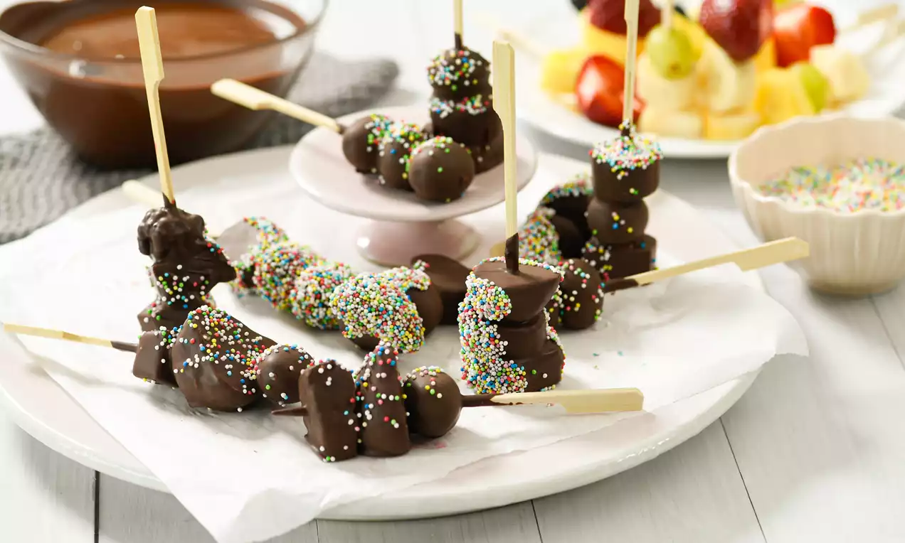 Drveni štapići s komadićima voća obloženim tamnom čokoladom, posuti sitnim šarenim mrvicama, složeni na bijelom tanjuru