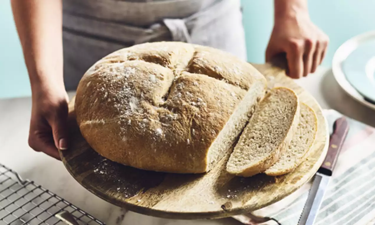 Rustic round loaf with a golden crust lightly dusted in flour, partially sliced to reveal a soft, airy crumb on a wooden board