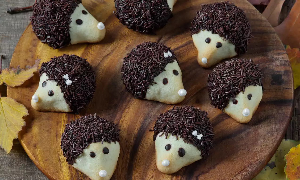 Malé světlé sušenkové ježky se zdobením z čokoládových drobků na dřevěném talíři, některé doplněny bílými cukrovými perličkami