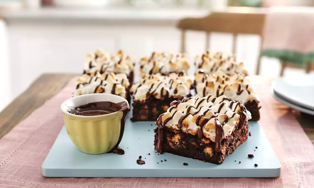 Smores brownies with rich chocolate layers, toasted marshmallows, and dark chocolate drizzle on a pastel blue board next to a green bowl of melted chocolate