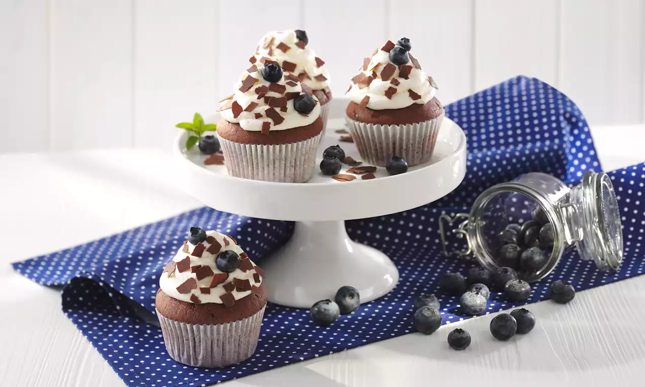 Blaubeer-Schokoladen-Muffins mit weißer Sahnehaube, Schokosplittern und Blaubeeren auf weißer Etagere auf gepunktetem blauem Tuch drapiert