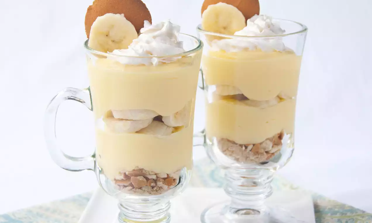 Two clear glass parfait cups layered with pale yellow banana pudding, banana slices, crunchy crumble, whipped cream, and cookie discs