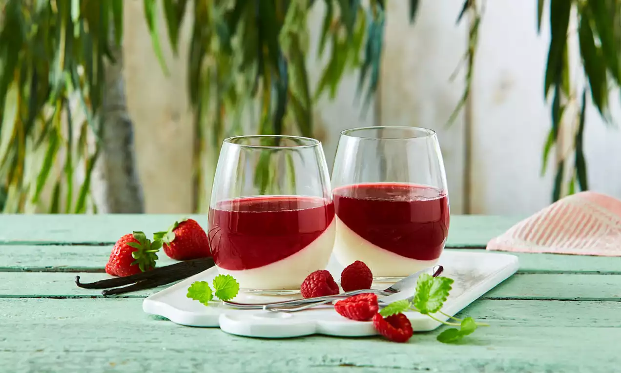 Panna cotta i glas med snehvid bund og glat hindbærrød gelé, pyntet med friske hindbær og grønne blade på en hvid bakke