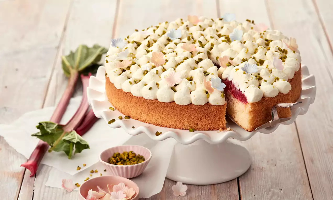 Rhabarber-Torte mit goldbraunem Boden, weißer Sahnehaube, Pistazien und Blüten dekoriert, auf einer gewellten Tortenplatte serviert