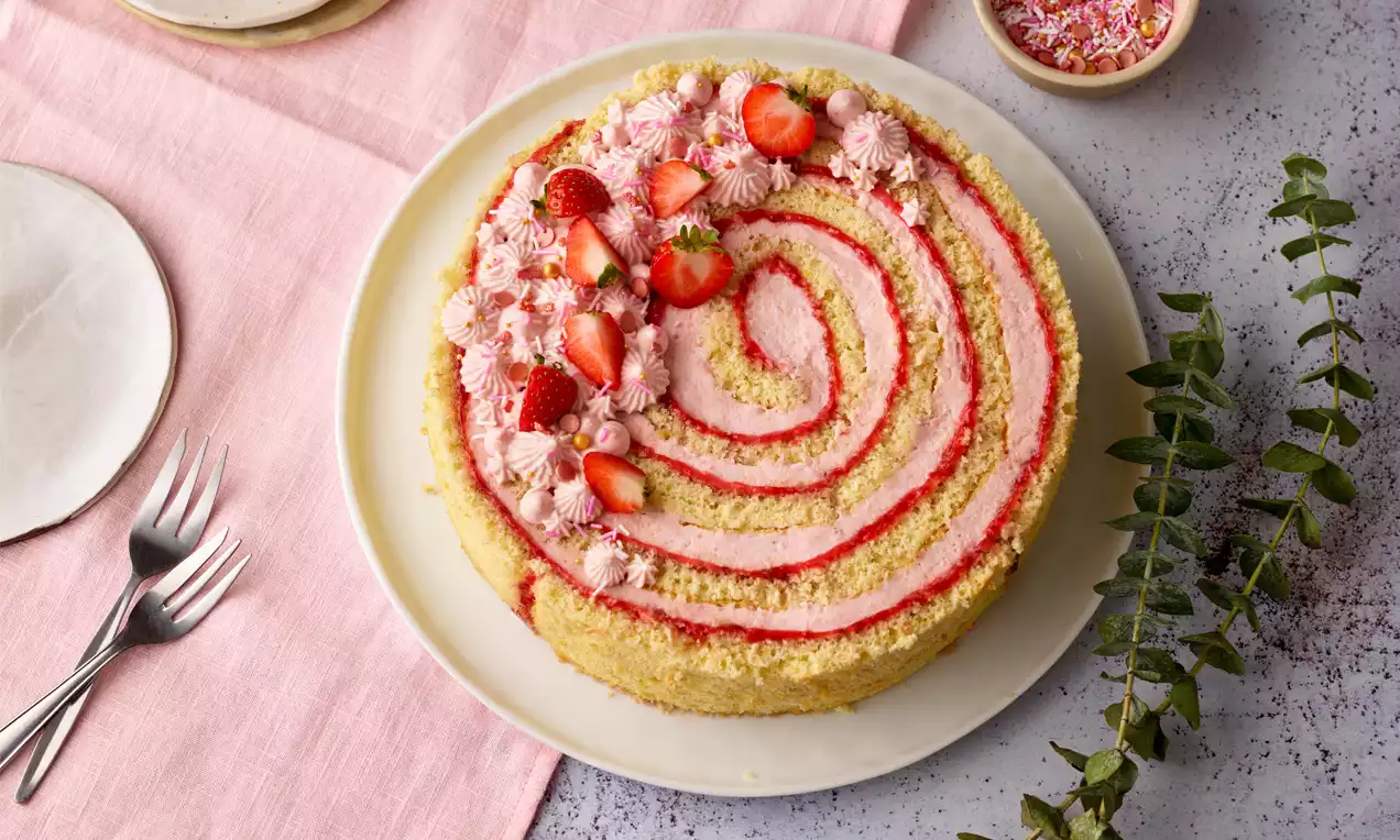 Wickeltorte mit hellgelbem Biskuit und spiralförmiger roter Füllung, dekoriert mit rosa Creme, frischen Erdbeeren und Sahnetupfen auf einem weißen Teller